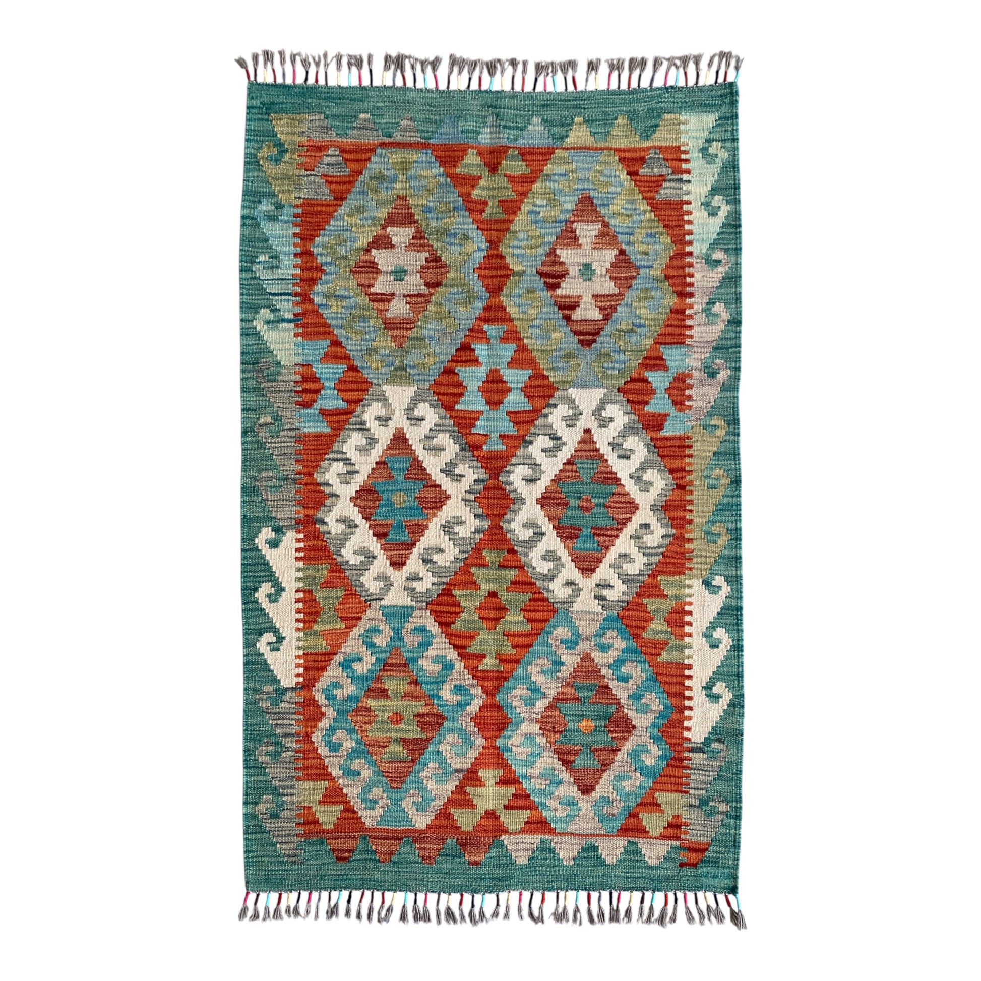 A rectangular kilim rug with a geometric diamond pattern in teal, rust red, cream, and light blue, finished with fringed tassels on the top and bottom.