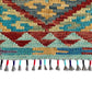 A close-up of a woven kilim rug showing a geometric pattern in red, blue,  teal, beige, and gray, finished with a fringe of multicolored tassels on a white background.