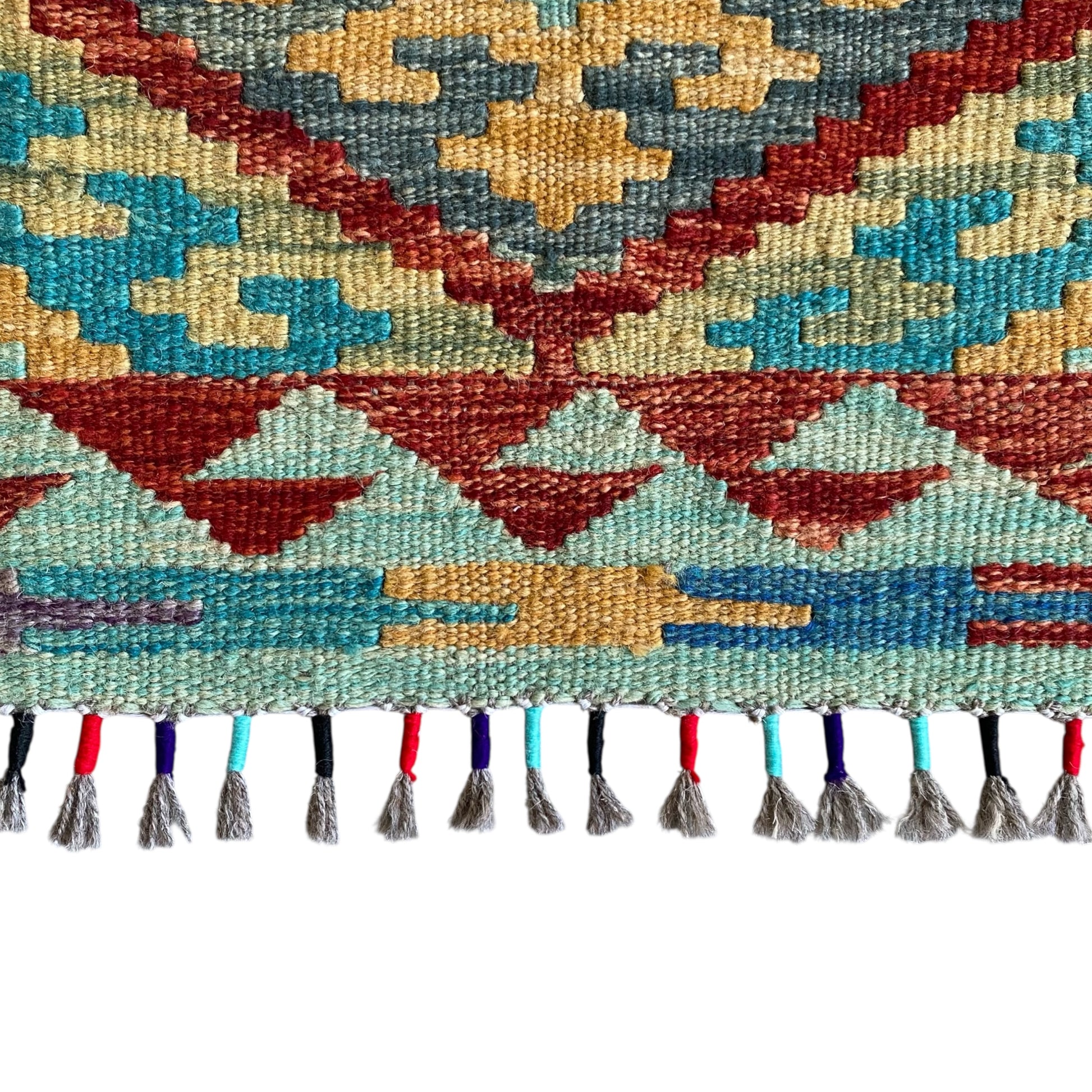 A close-up of a woven kilim rug showing a geometric pattern in red, blue,  teal, beige, and gray, finished with a fringe of multicolored tassels on a white background.