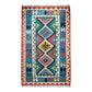 A hand woven area kelim rug with a vibrant Turkish Nomadic geometric pattern, featuring a central diamond motif in pink, yellow, blue, and black on a cream background, surrounded by a red and blue border with fringe on the top and bottom.